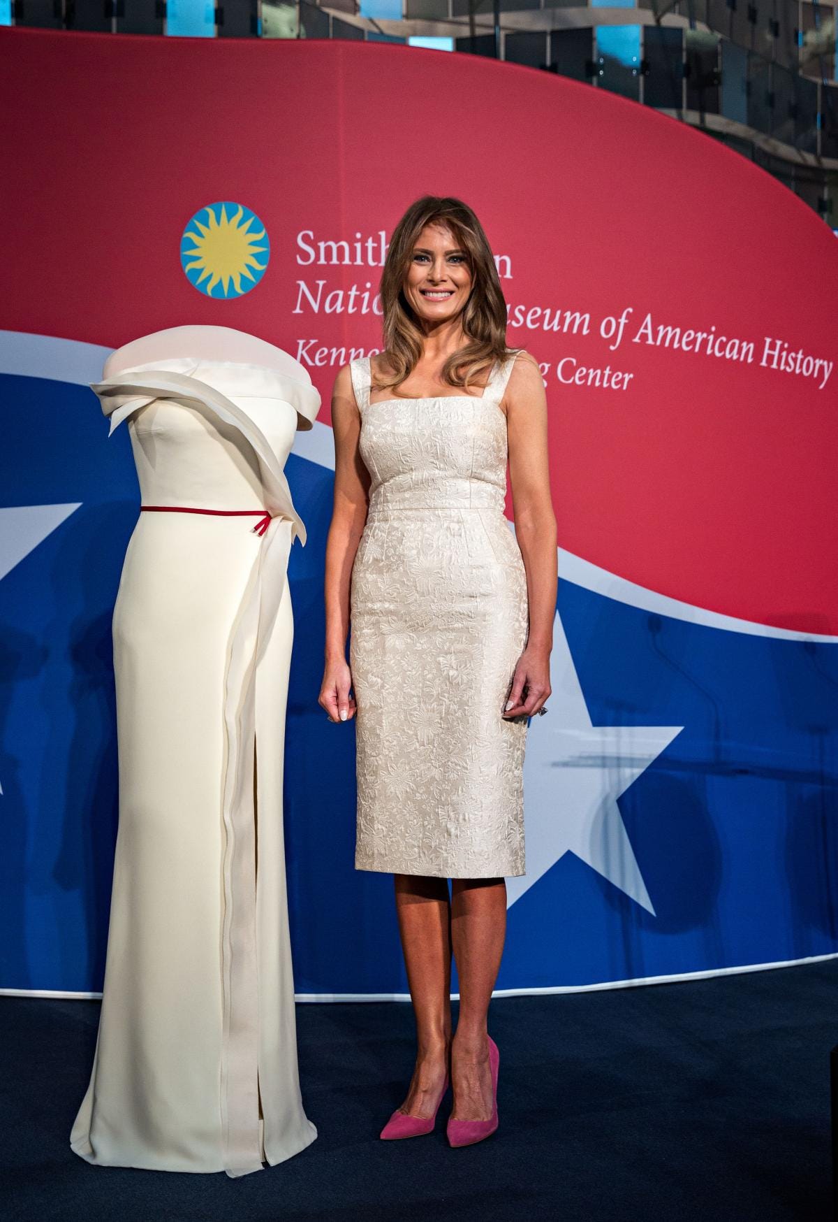 Posted photo on stage. On left, white gown on a form. On right, First Lady Melania Trump smiling beside her gown.