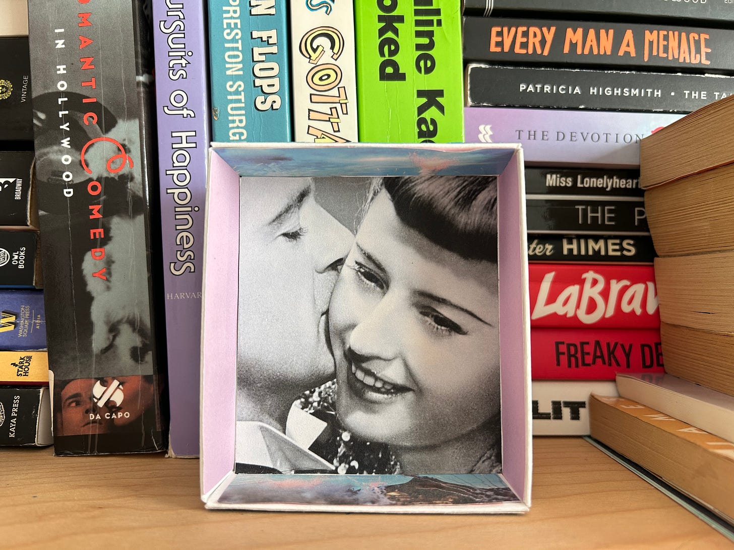 Image shows a black and white, close-up photograph of a couple pasted to the bottom of a paper box, which is rested on one side and facing outward, giving it the appearance of a frame. In the photograph, the man on the left kisses the cheek of the woman on the right. The framed photograph is set against a shelf of colorful books, whose spines are facing out. 