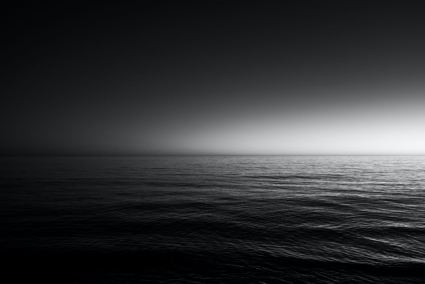 A black and white photo of a body of water with gentle waves rippling over the top. The sky is dark with a little bit of light on the horizon. The water is also dark.