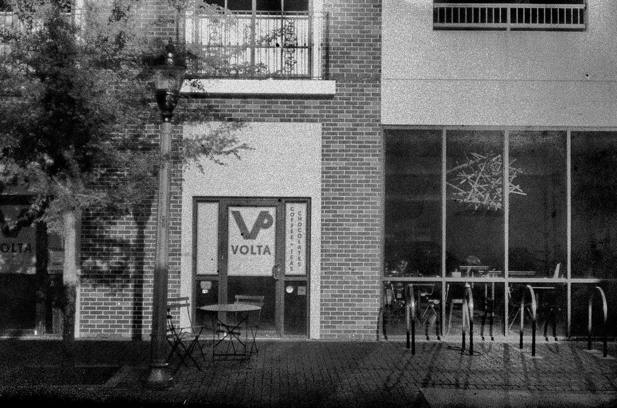 A black and white photo of the outside of Volta coffee shop with a tree, a lamp post, and a small table with two chairs on the sidewalk. 