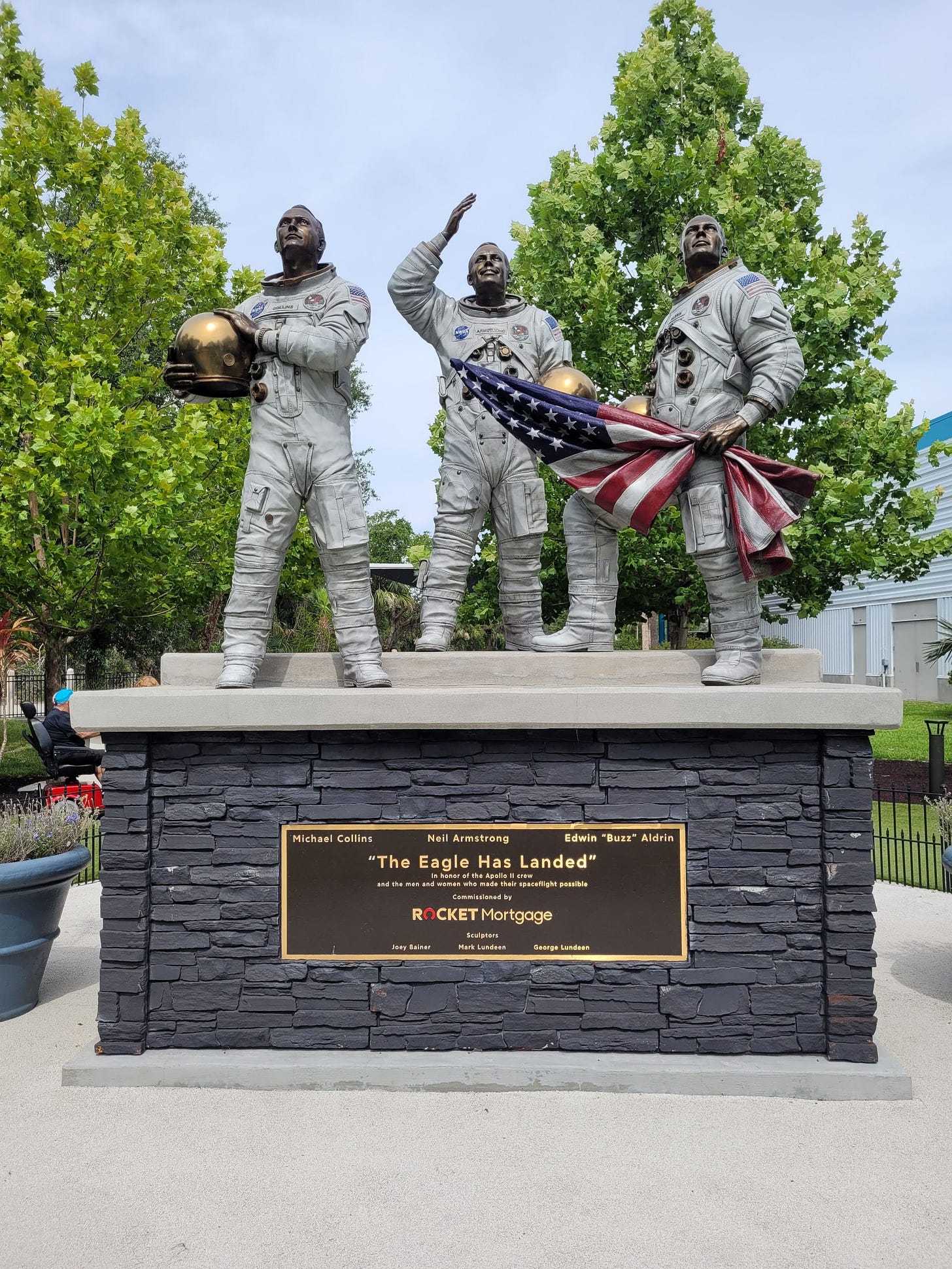 Um pedestal de tijolos acinzentados e em cima as esculturas dos três astronautas da Apollo 11. Todos com o capacete na mão, Neil acenando e Buzz segurando a bandeira dos EUA. Ao fundo dá pra ver um céu azul e árvores.