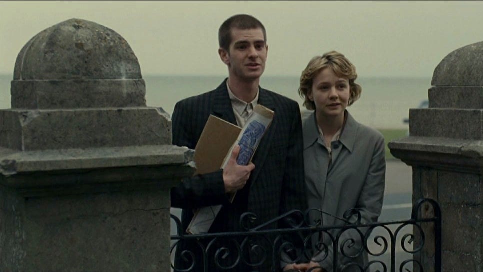 Still from Never Let Me Go that shows Tommy and Kathy standing at a gate, Tommy clutching a bunch of rolled papers and notebooks of art