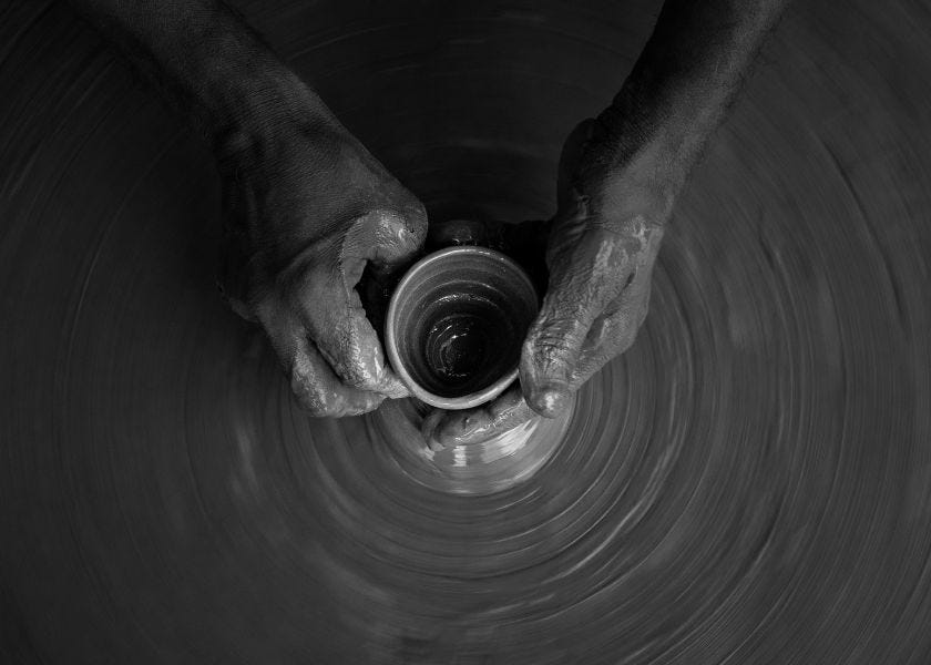 The image shows a pair of hands skillfully shaping a small clay pot on a spinning pottery wheel. The hands are coated with clay, indicating the ongoing process of molding the pot. The background and the wheel appear blurred due to the motion, emphasizing the focus on the hands and the delicate work being done. The image is in black and white, giving it a timeless and artistic feel.