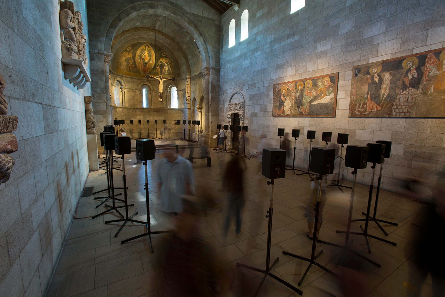 Photograph of a medieval stone chapel, with 40 speakers dispersed throughout the space as a handful of people move between speakers