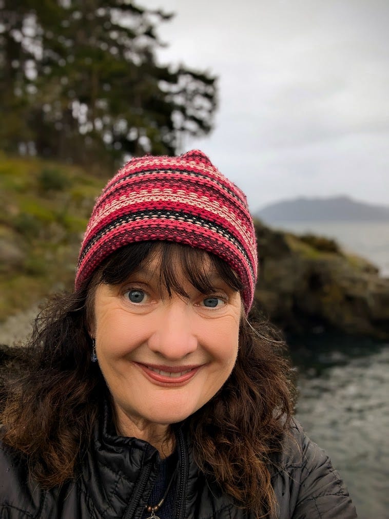 Brown-haired woman in pink and red striped hat smiling with tall trees and bay water in background. 