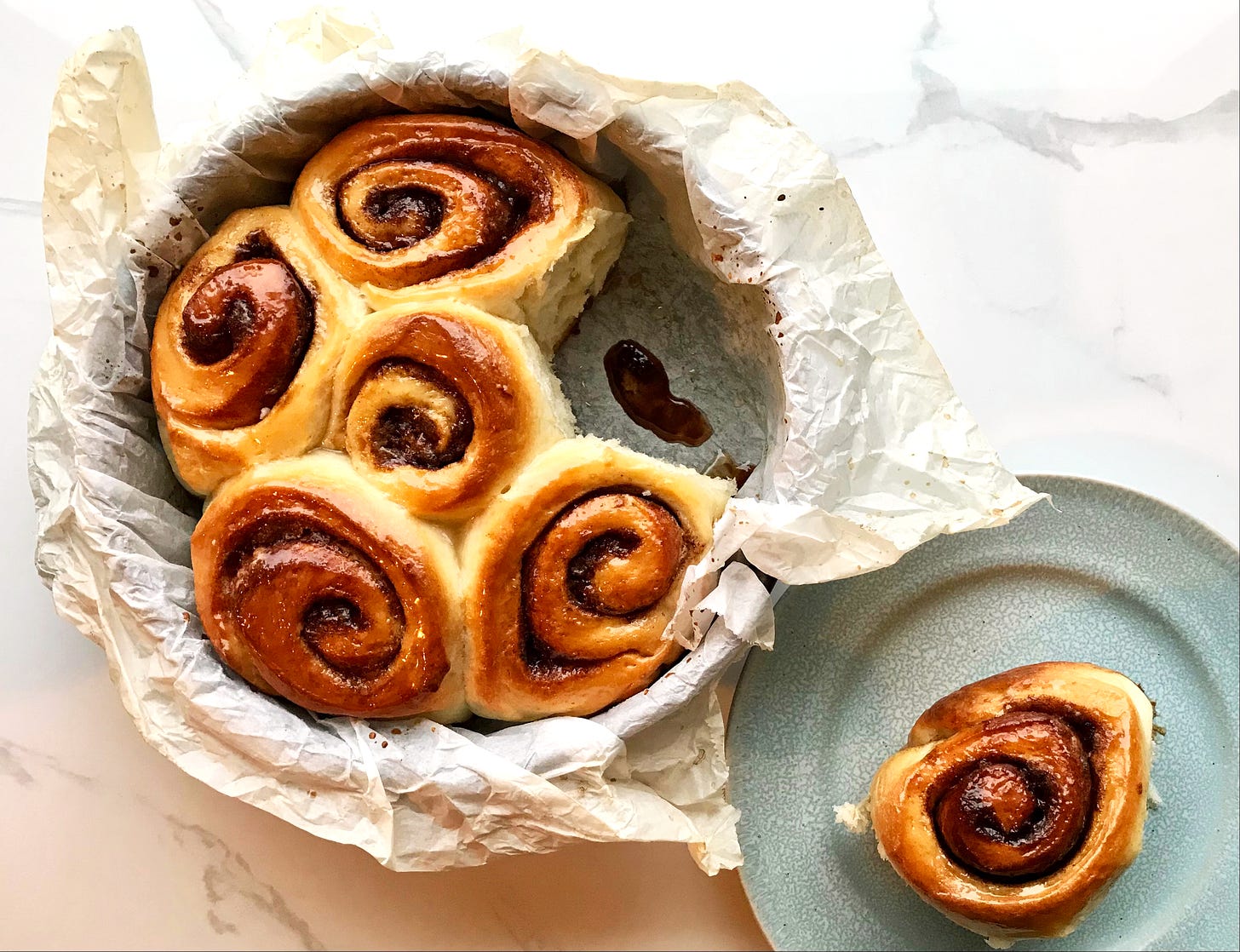 A parchment-lined cake tin with five baked and glazed cinnamon rolls. In the gap where one is missing is a puddle of melted cinnamon, sugar and butter. Beside the tin is a plate with the sixth cinnamon bun.