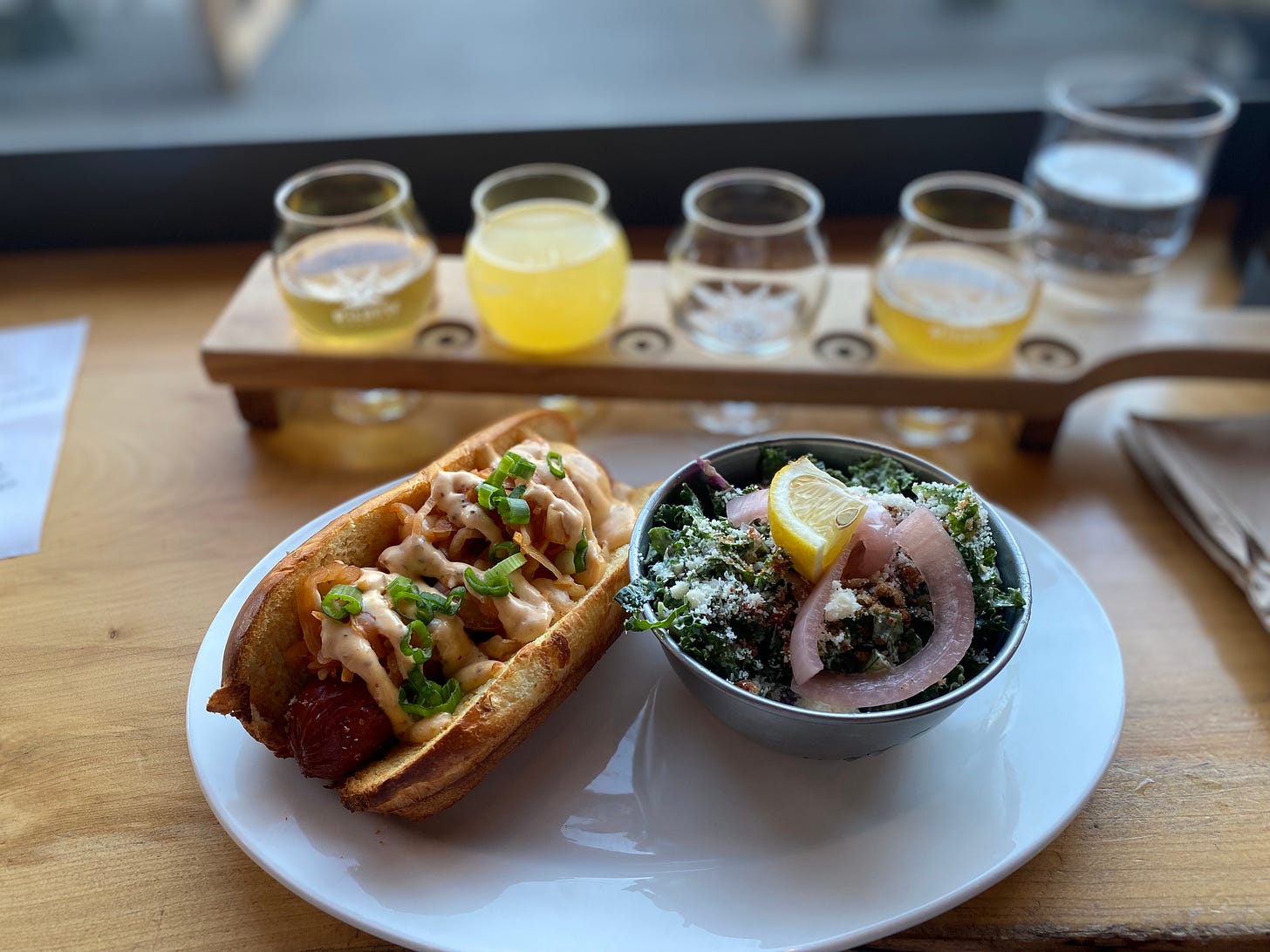 A hot dog with kimchi, spicy mayo, and green onions on a white plate next to a small metal bowl of kale caesar salad with pickled onions, dusted with breadcrumbs and parmesan and garnished with lemon. In the background is a flight of beer in a wood paddle.