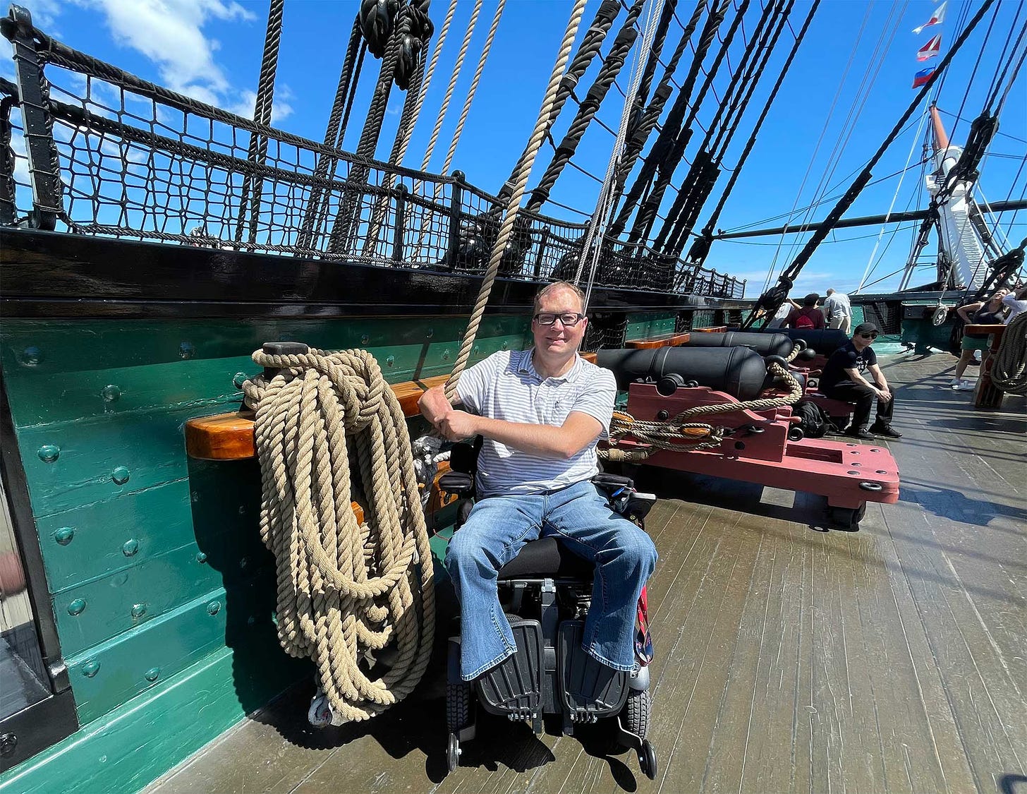 John seated in his wheelchair on the wooden deck of the Constitution with a cannon in the background.
