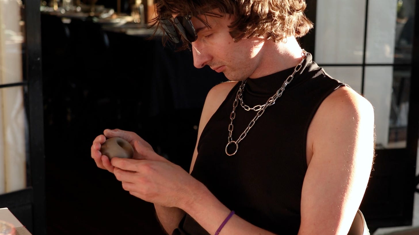 Image description: Ari moulds clay in his hands. He is wearing black sunglasses on his forehead, a black singlet and silver chain necklaces.