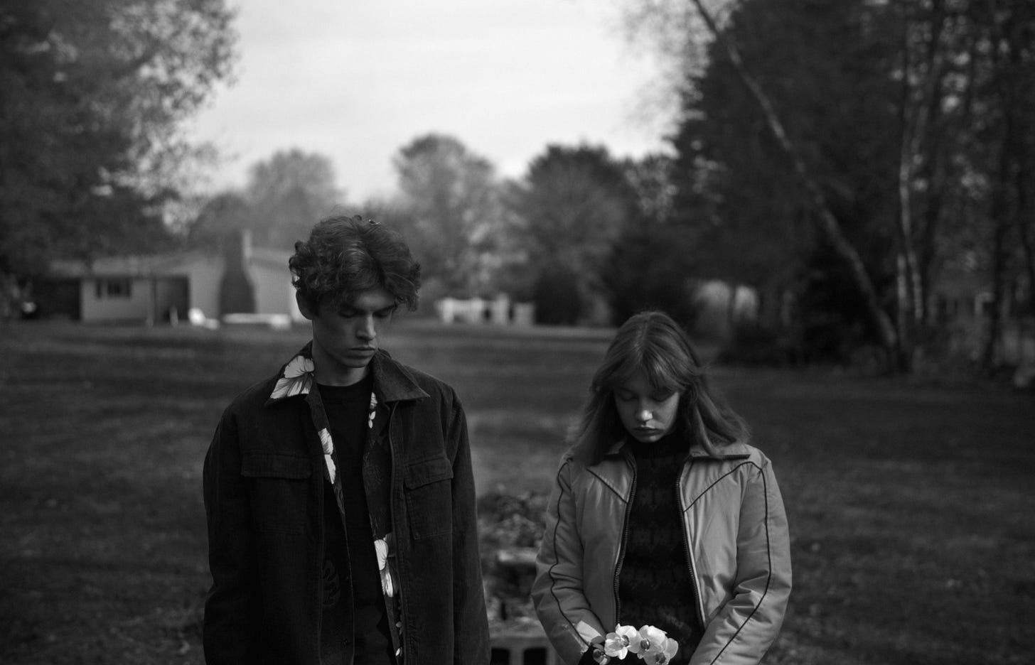 A black and white photo of two individuals outdoors, looking down, one holding flowers.