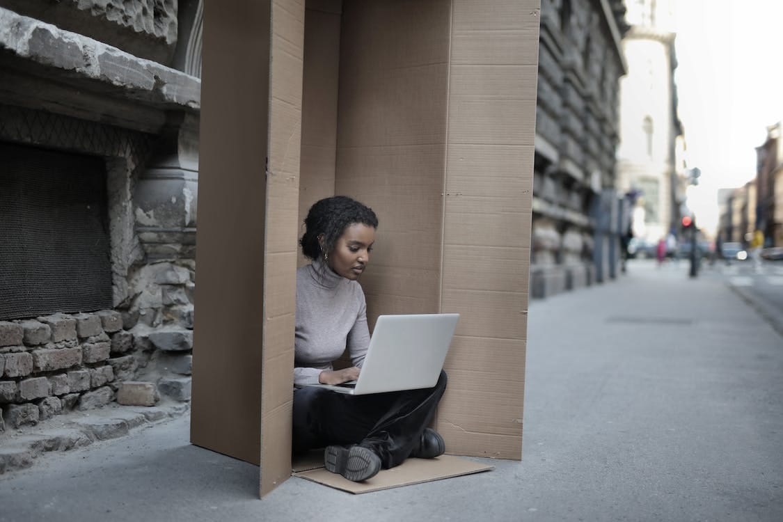 Free Young female introvert in casual clothes typing on netbook while sitting with crossed legs inside cardboard box located on asphalt road in city in afternoon Stock Photo