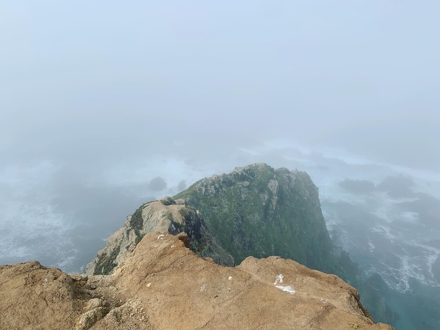 An orange rock peninsula jutting out into a foggy sea with waves crashing