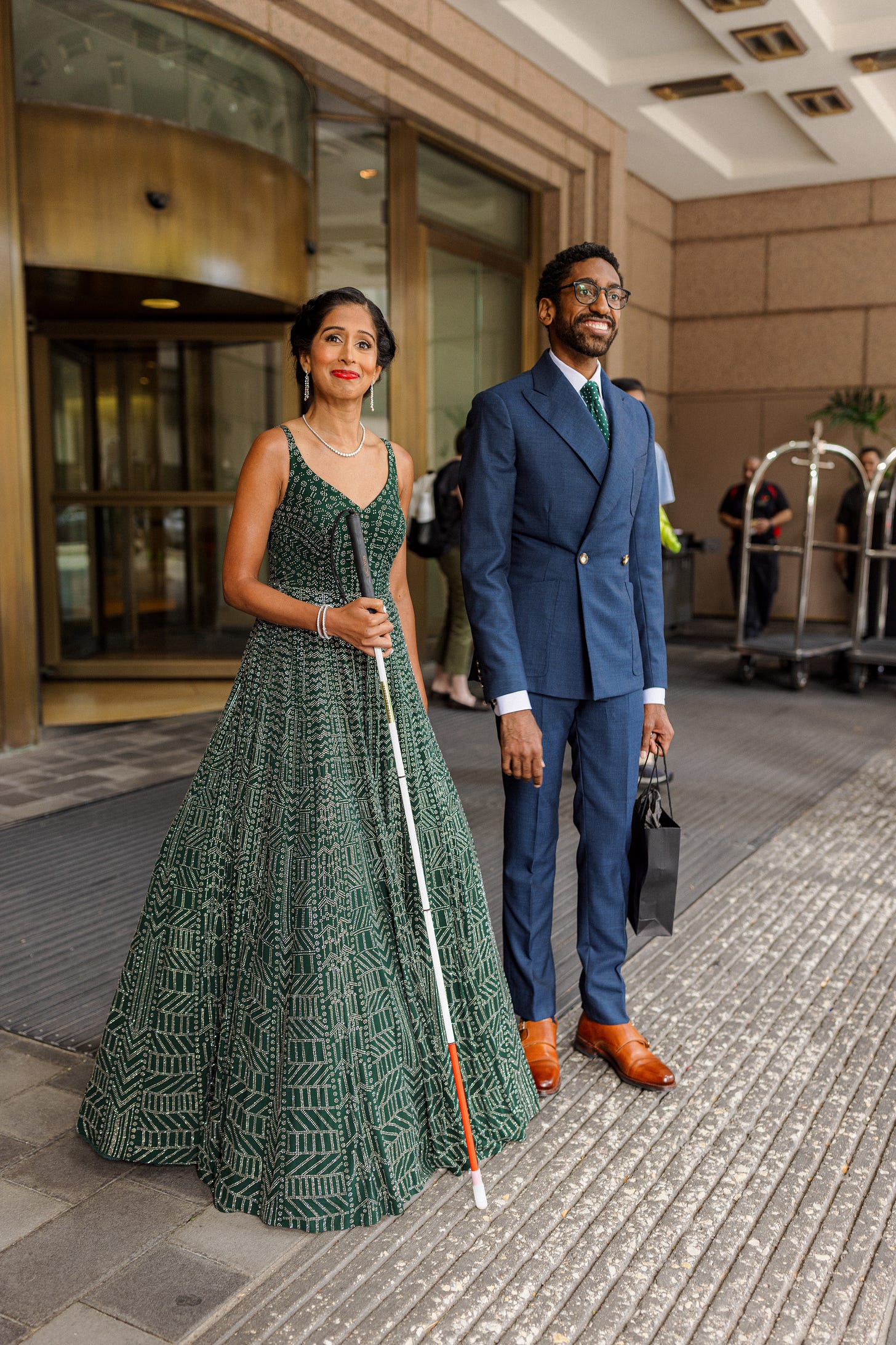 The couple stands in front of the Fairmont, about to leave for the ceremony. Qudsiya, wearing a stunning, bottle green floor length gown covered in beads, holds her white cane in front of her. Her smile, with lips closed, is full of anticipation but measured. Sean, in a navy blue double breasted suit, holds a small black gift bag in his left hand and looks off, smiling, toward where family members are arriving. Little pops of color stand out: Qudsiya, with her gleaming diamond jewelry and the white of her cane, her bold red lipstick and the red stripe near the tip of her cane; Sean with his white shirt collar and cuffs.
