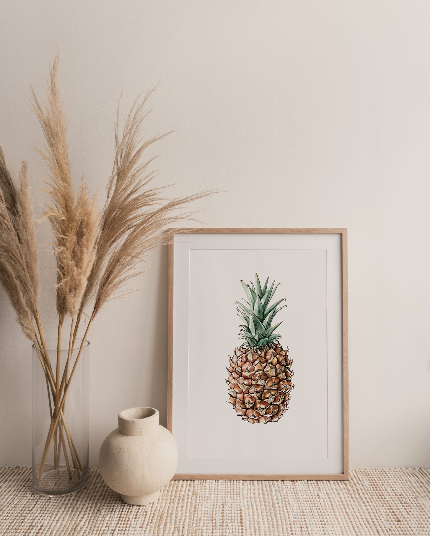 A framed print of a pineapple painted in watercolor, sitting on a table with a vase of tall grasses to the left of it 