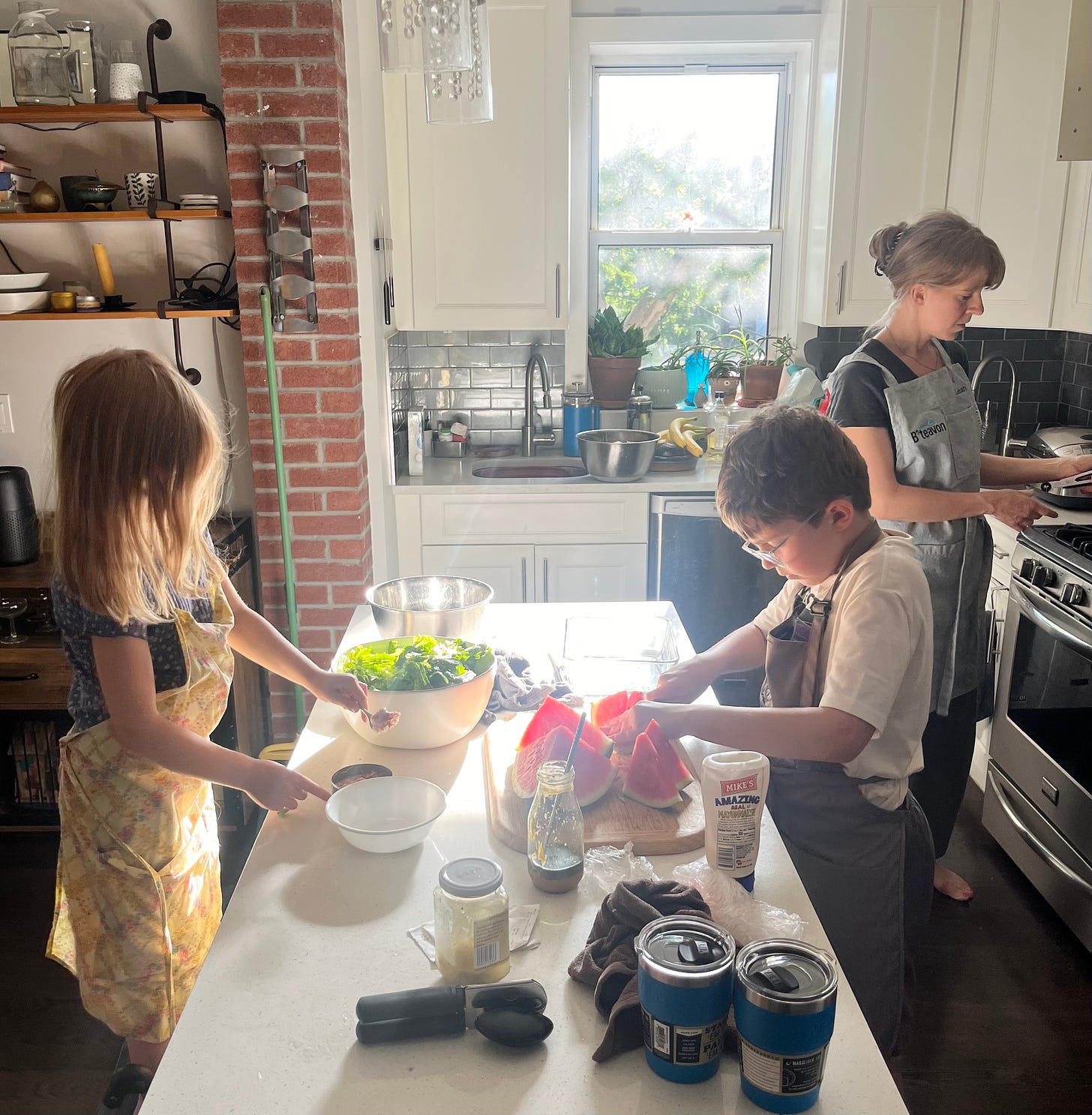 A young girl and young boy stand at a kitchen counter preparing a salad and slicing watermelon, while their mom stands in the background in front of the oven.