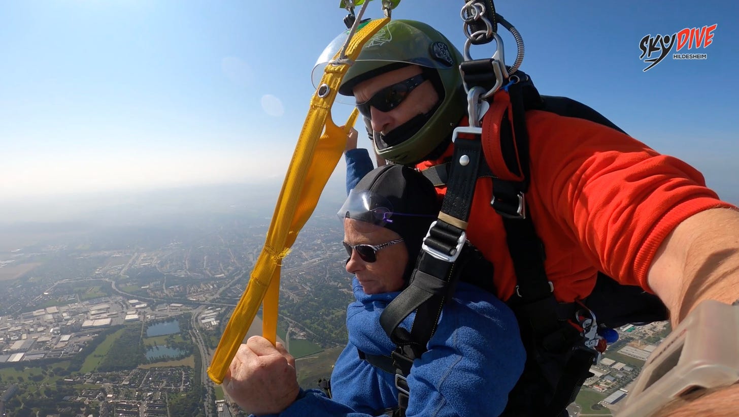 A tandem skydiver wearing a blue fleece and sunglasses is harnessed with an instructor in a green helmet and orange shirt. They are gliding under a parachute high above Hildesheim, Germany, with a clear blue sky and city landscape below. This moment captures the essence of releasing control and embracing freedom.