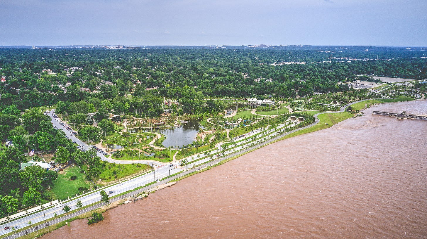 Seen from above, the muddy waters of the flooded Arkansas River reach almost to the River Parks trail at Gathering Place: a large park with carnival-like structures, an irregularly shaped pond, and winding paths around extensive landscaping. The backdrop is a lush, expansive view of midtown and south Tulsa below a slightly hazy, cornflower blue sky. Inside Gathering Place, the parking lot appears full.