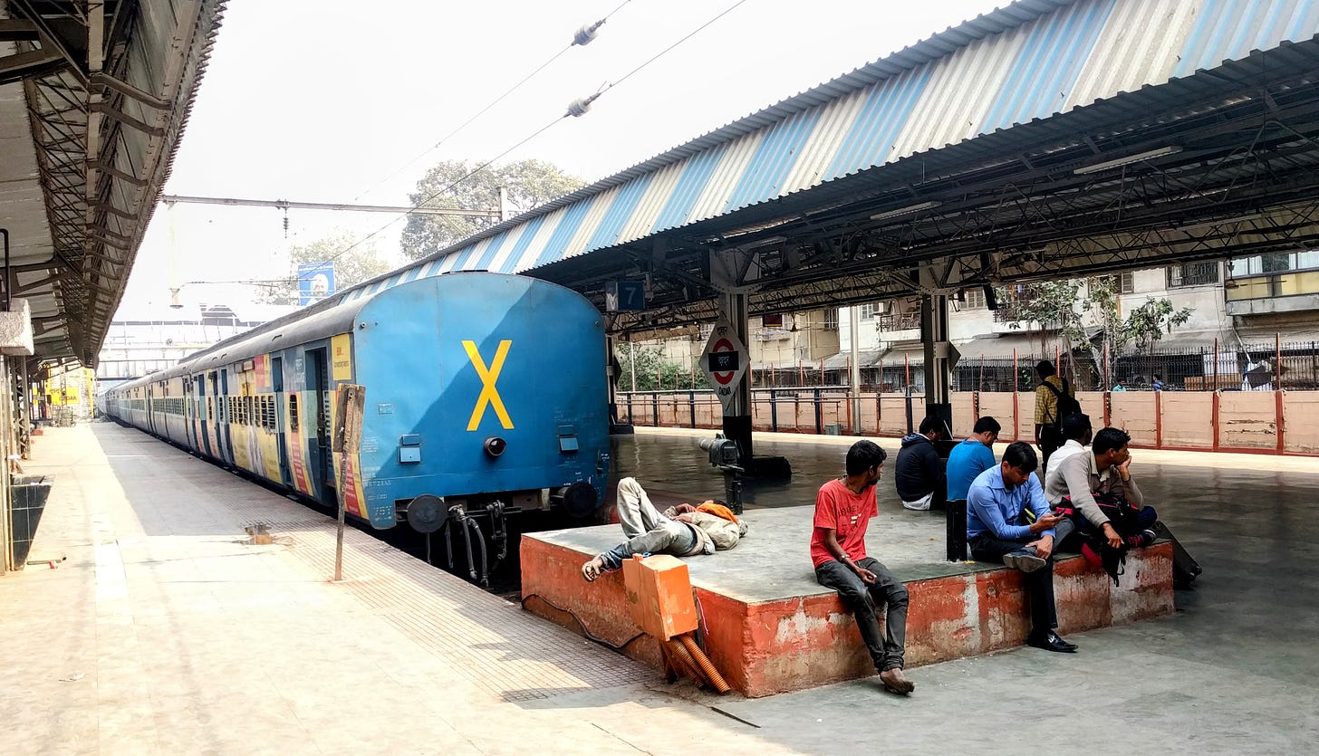 Indian men on a train platform check their phones and nap. A colorful train with a big yellow X on the end sits on the track behind them.