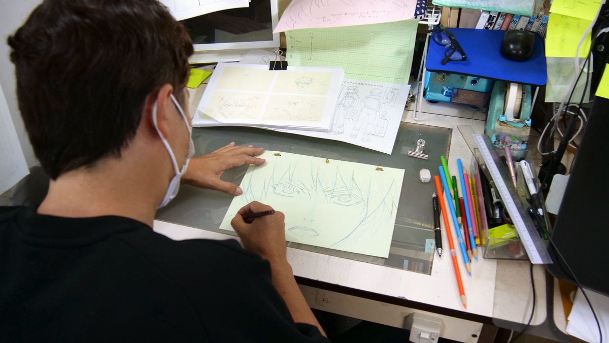 Photograph of a man at his desk drawing an anime character on paper, taken from a high vantage point looking down from behind him. Additional sheets of paper, drawing pencils, and other stationery are scattered on the desk.