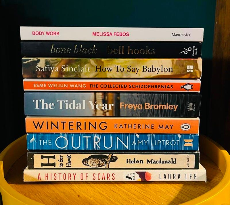 A pile of paperback books on a wooden yellow table. The books are from top to bottom: Body Work by Melissa Febos, bone black by bell hooks, How To Say Babylon by Safiya Sinclair, The Collected Schizophrenias by Esme Weijun Wang, The Tidal Year by Freya Bromley, Wintering by Katherine May, The Outrun by Amy Liptrot, H is for Hawk by Helen Macdonald and A History of Scars by Laura Lee