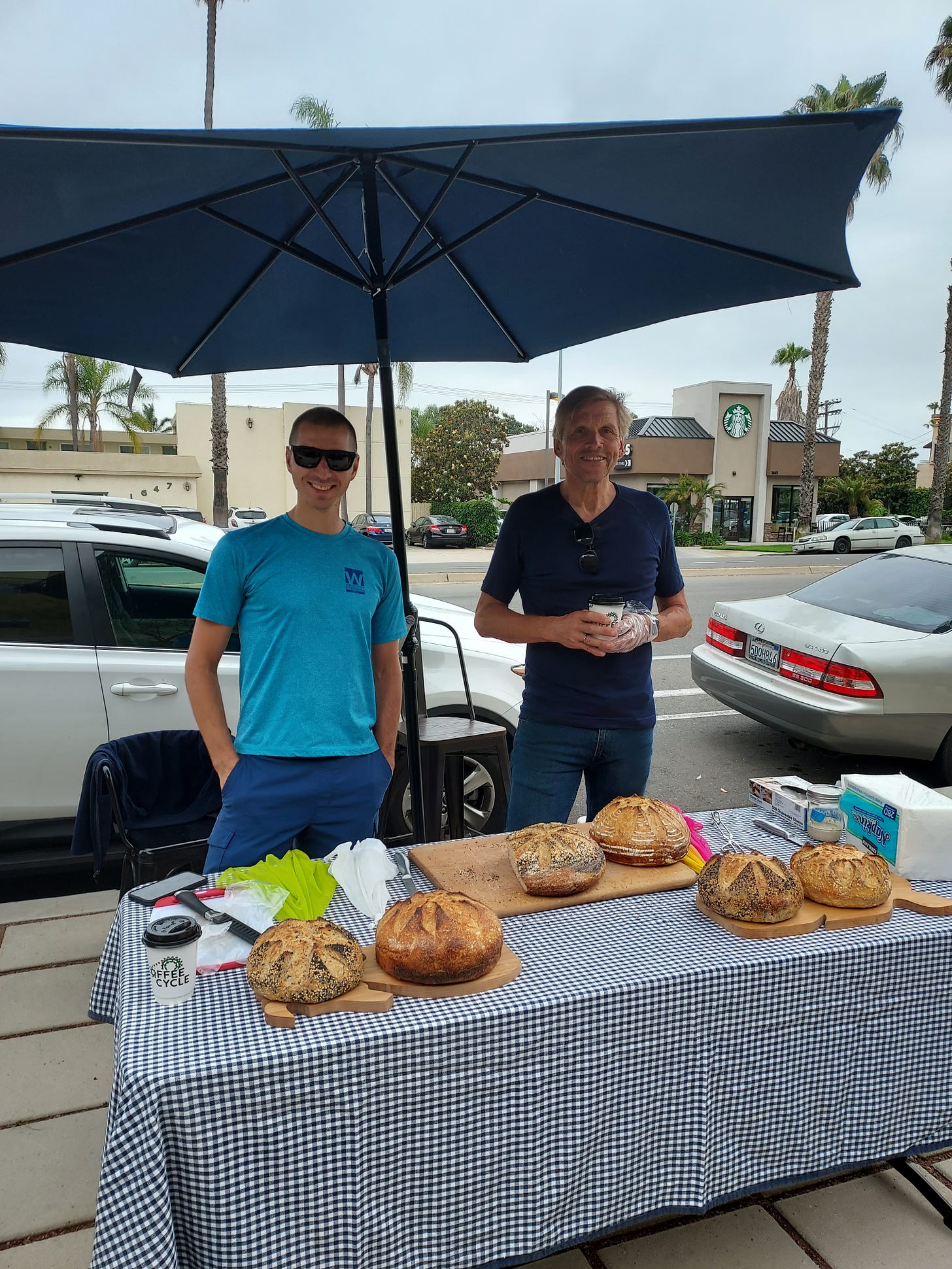 Two men stand behind a table filled with sourdough bread under a blue umbrella at a sidewalk maker's market.