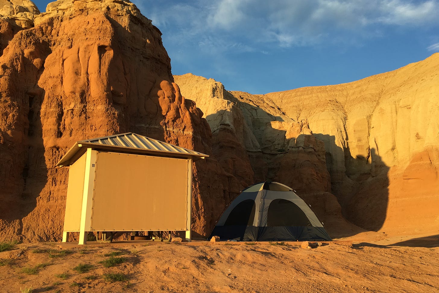 A blue dome tent hides in the shade of a picnic table shelter with a backdrop of Goblin Valley State Park hoodoos.