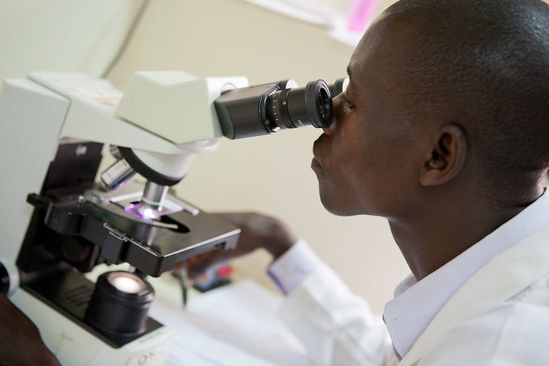 An African man wearing a lab coat and looking through a microscope.