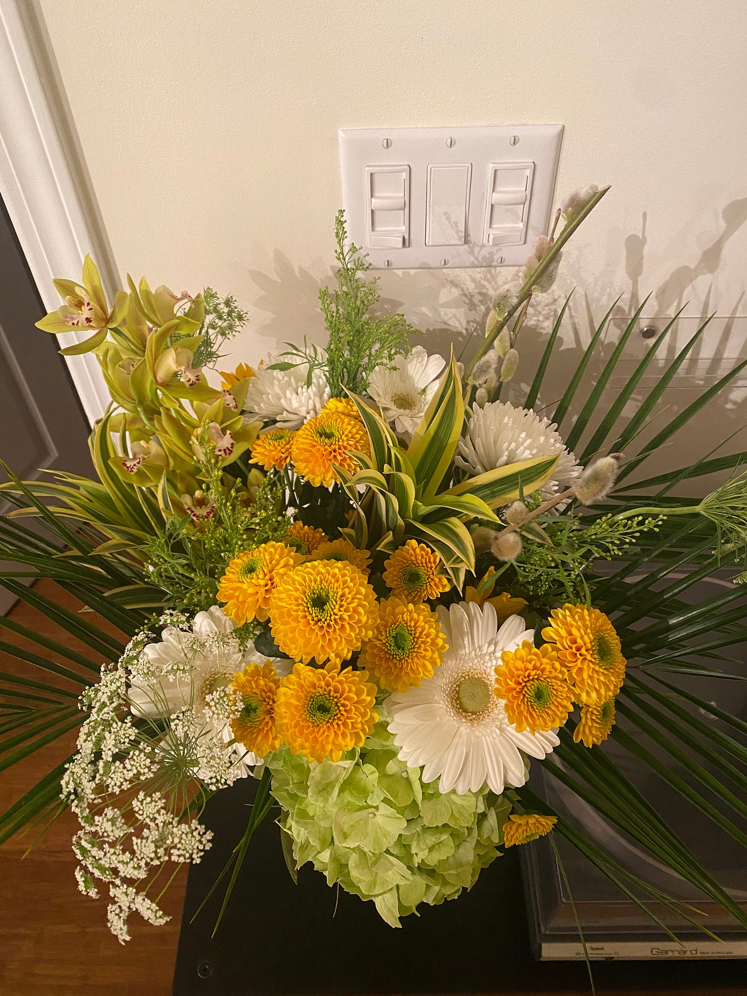 On a black shelf next to a record player, a cylindrical glass vase of flowers, mostly yellows and whites with tropical-looking greens mixed in. 