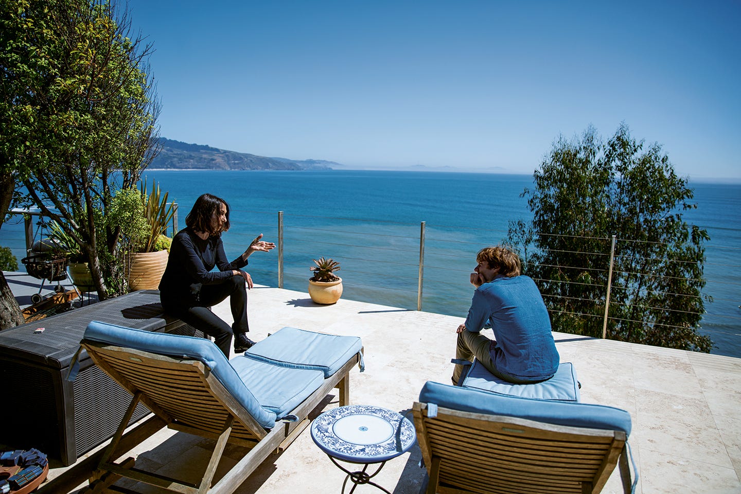 Suzanne Ciani and Spencer Tweedy sit on patio furniture on a concrete patio overlooking the Pacific ocean.