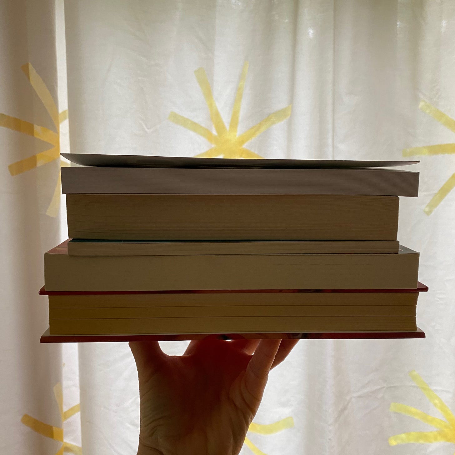 I am holding up five books, spines hidden, against a white curtain patterned with yellow suns.