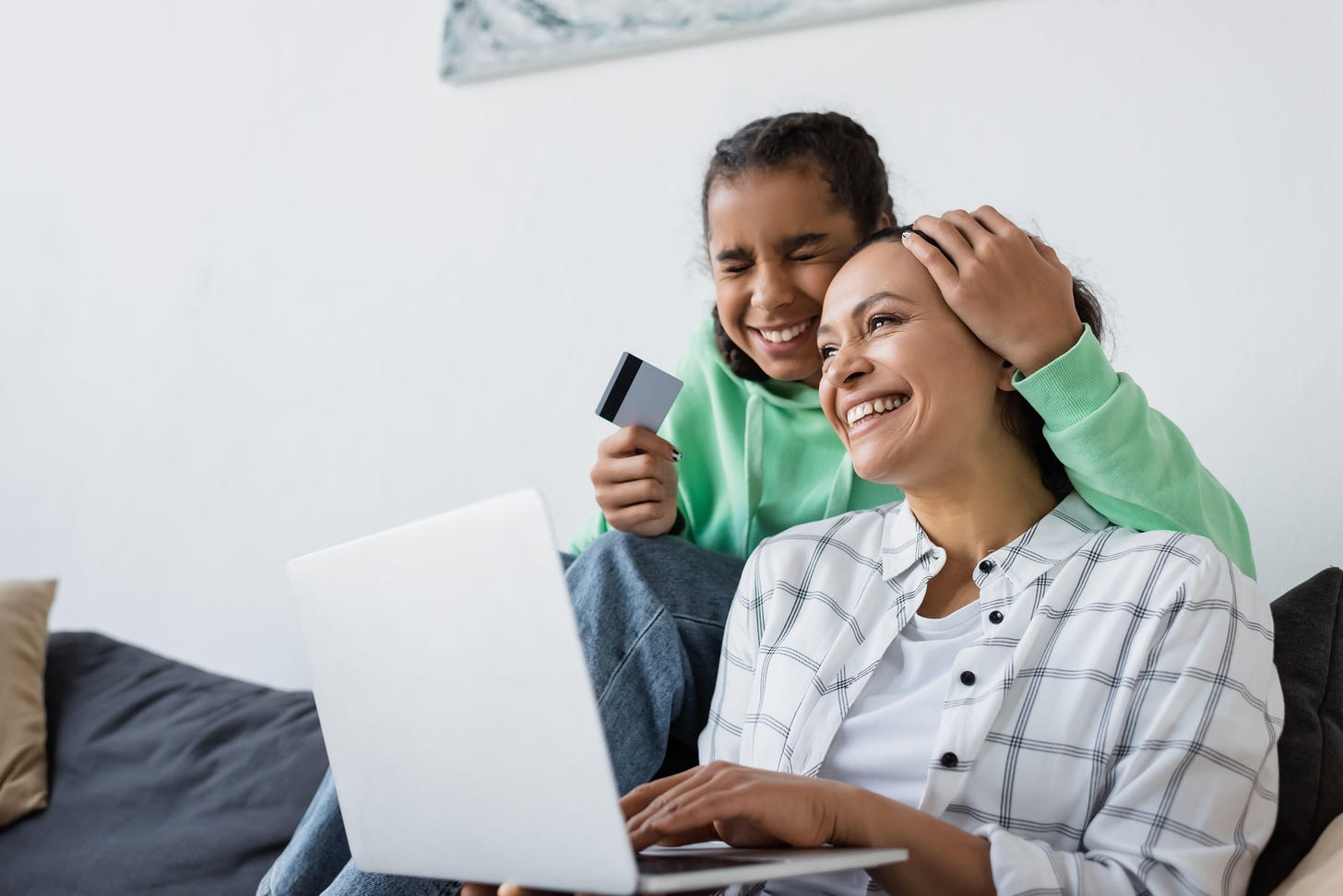 excited african american teen girl with closed eyes girl holding credit card near smiling mom and laptop