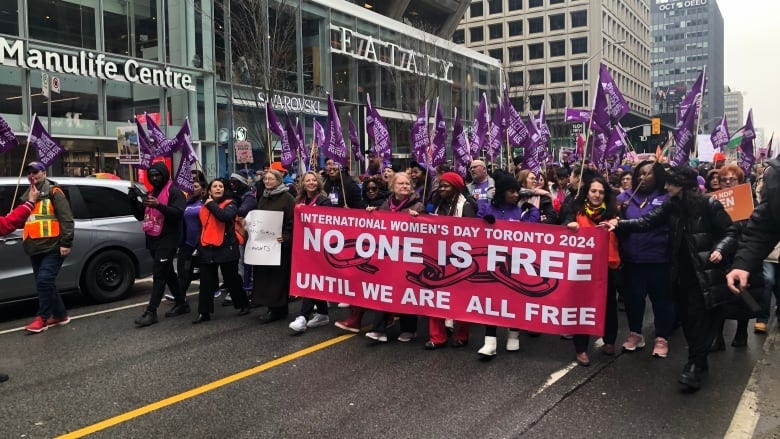 Protestors march down a street waving flags and carrying a banner that reads: "International Women's Day Toronto 2024 NO ONE IS FREE UNTIL WE ARE ALL FREE"