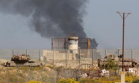 Black smoke rises behind the border fence; armoured vehicles and what may be a watchtower and agricultural equipment are in the foreground