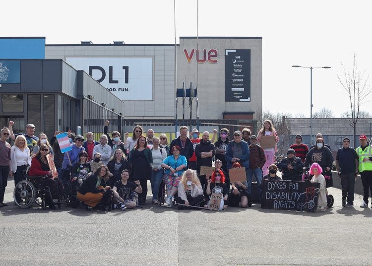A photograph of a group of 30-40 people gathered to protest against welfare cuts. They are in a car park and some are in wheelchairs, others hold placards. All face the camera.