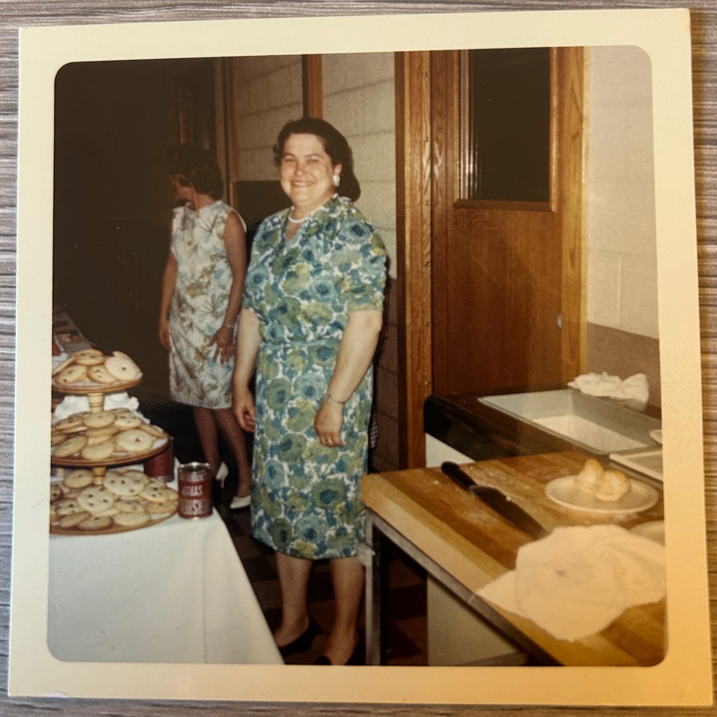 A smiling woman in a blue flowered dress stands in front of pastries. There's another woman in the background looking off camera. The colors of the photo are faded.