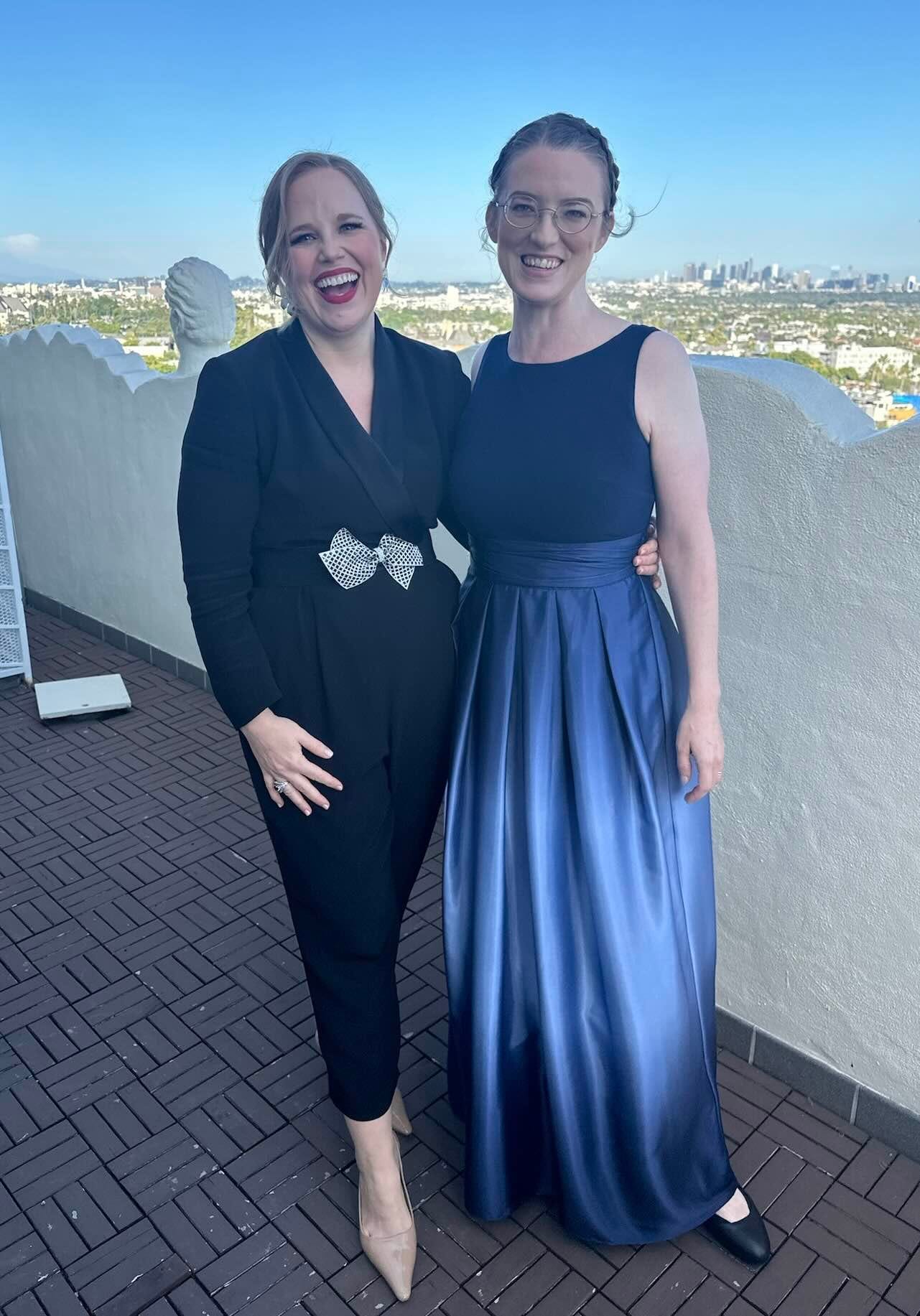 Lauren and Jodi on a terrace overlooking LA. Lauren is wearing a black tux jumpsuit with a silver bow belt; Jodi is wearing a blue gown. 