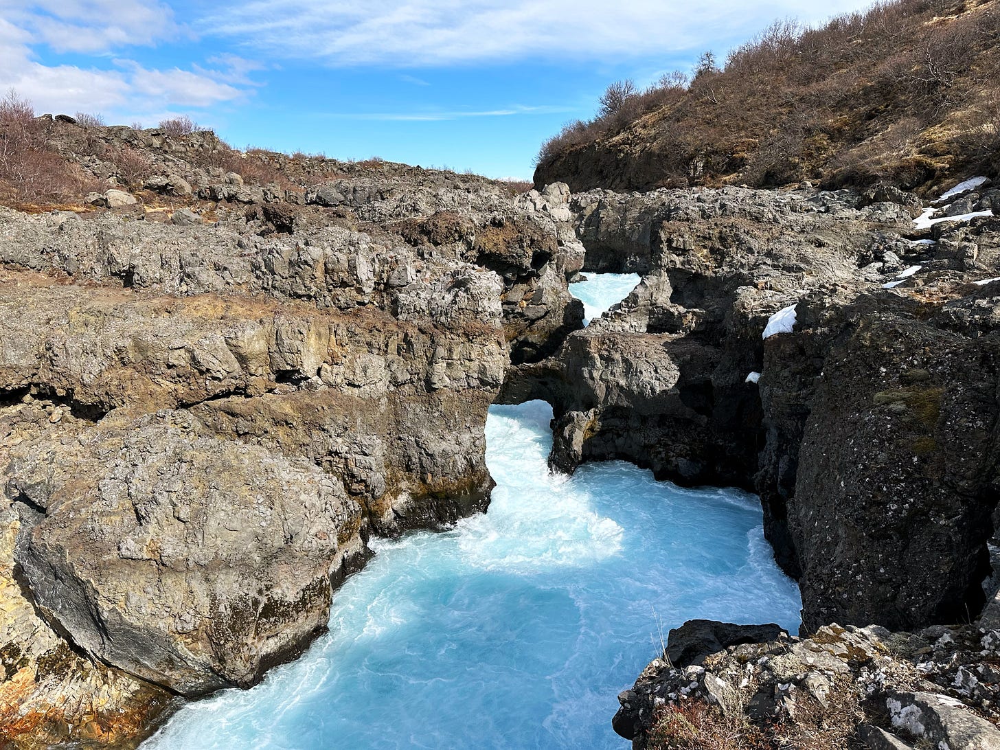 Foamy, light blue water flows downhill in a series of rapids to the river below.  The rocks are black and gray lava rock and the sky is bright blue with wispy white clouds.