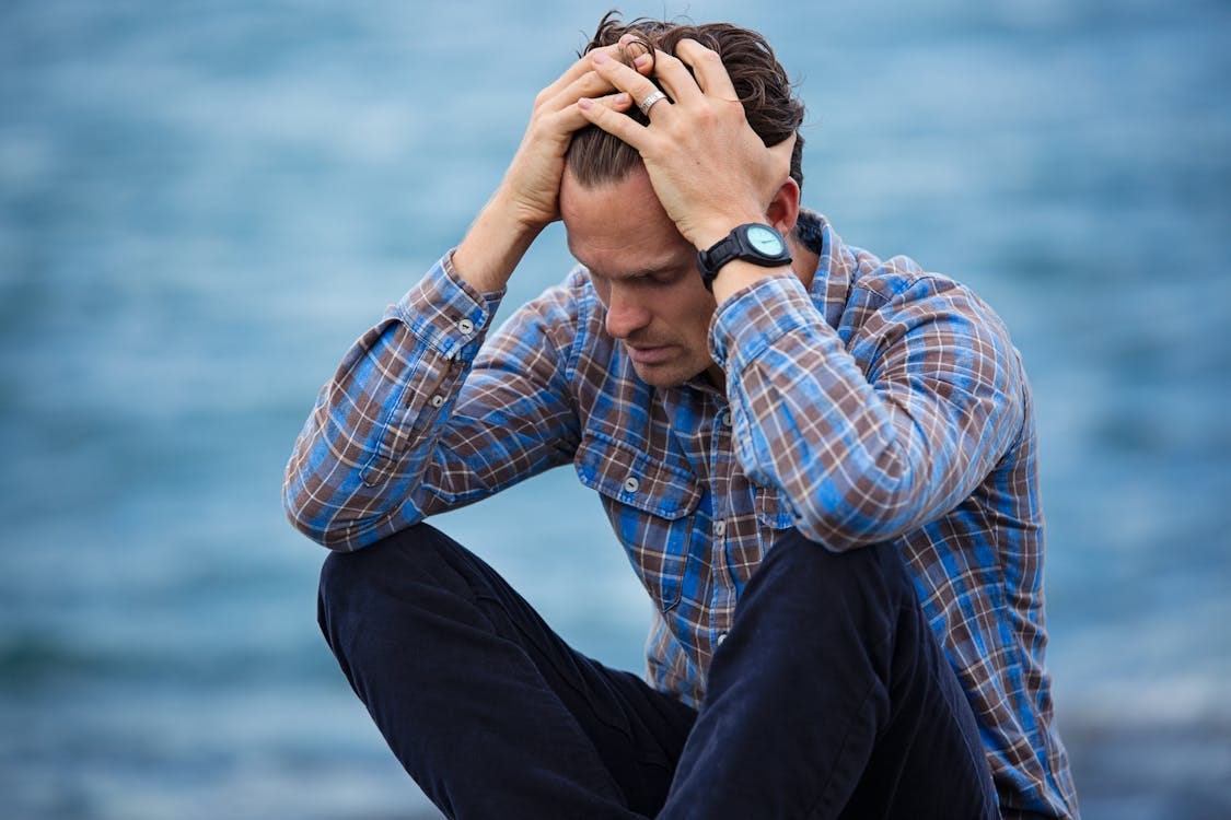 Free A man in a plaid shirt sits by the water looking distressed, symbolizing stress. Stock Photo
