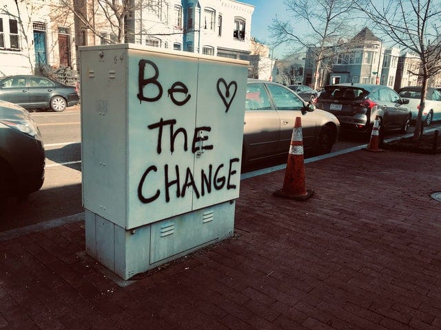 Photo looking towards the road, of a white electrical box on the  side of a red brick paved sidewalk, spray-painted with black lettering "Be the change" and a heart. Two orange traffic cones are nearby, in front of parked cars.