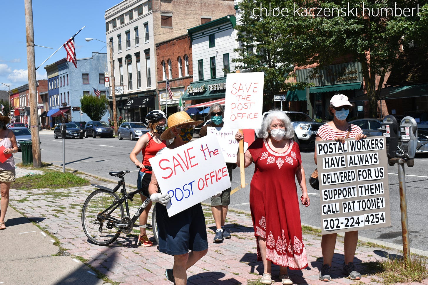 demonstrators holding signs that say save the post office, standing on a sidewalk of a main road in a small town on a sunny day