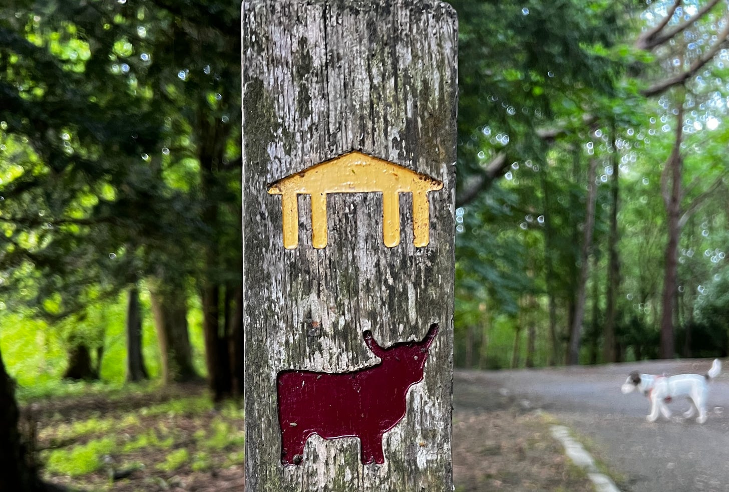 Post with two icons. One of a building with columns, the other of a highland cow. A blurred Jack Russel in the background to the right of the post looks like it's talking to the cow