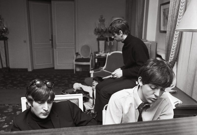 John and Paul deep in thought as they compose at the piano in their suite at the George V Hotel with George and Ringo in the background. Paris, January 1964.