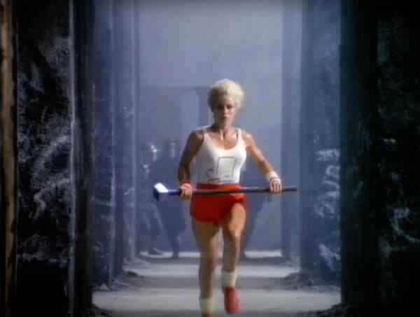 A woman with short white-blonde hair, a white tank top and red shorts runs through a corridor with a sledgehammer