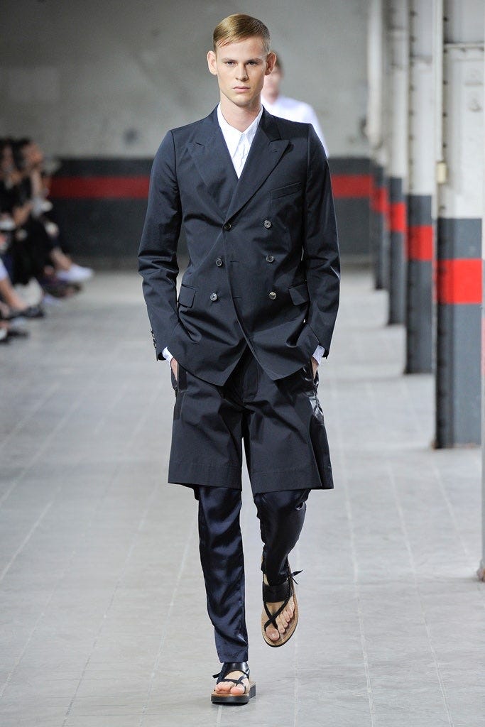 A model on a fashion catwalk, who is a white man with blonde hair, wearing a navy double breasted jacket and white shirt with slim navy trousers layered under wide legged navy shorts and black sandals with a chunky sole. A model on a fashion catwalk, who is a white man with blonde hair, wearing a navy double breasted jacket and white shirt with slim navy trousers layered under wide legged navy shorts and black sandals with a chunky sole.