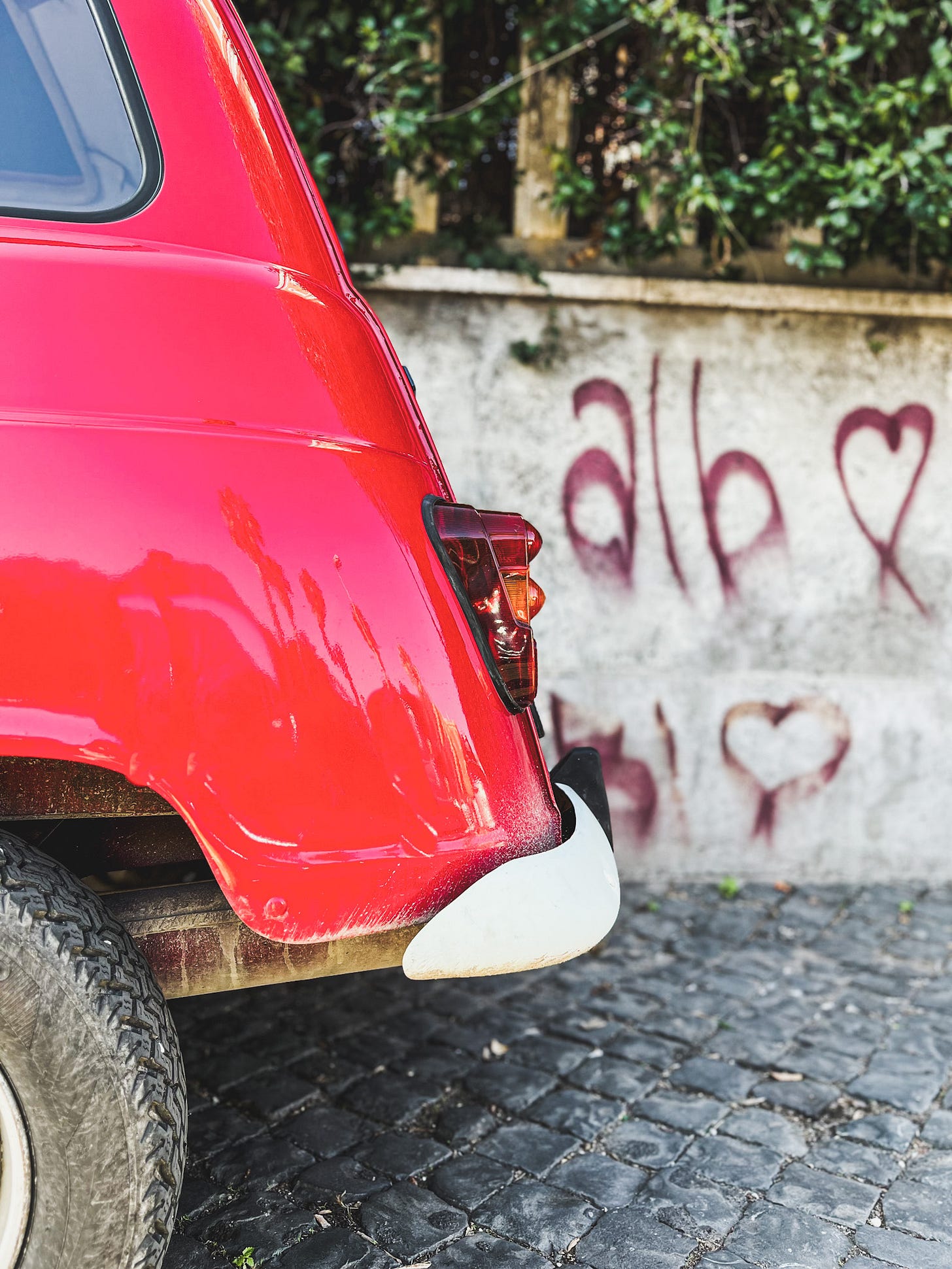 Rear of a vintage red Renault parked on cobblestone with heart graffiti on the wall behind it. Photo by Michelle Ciani