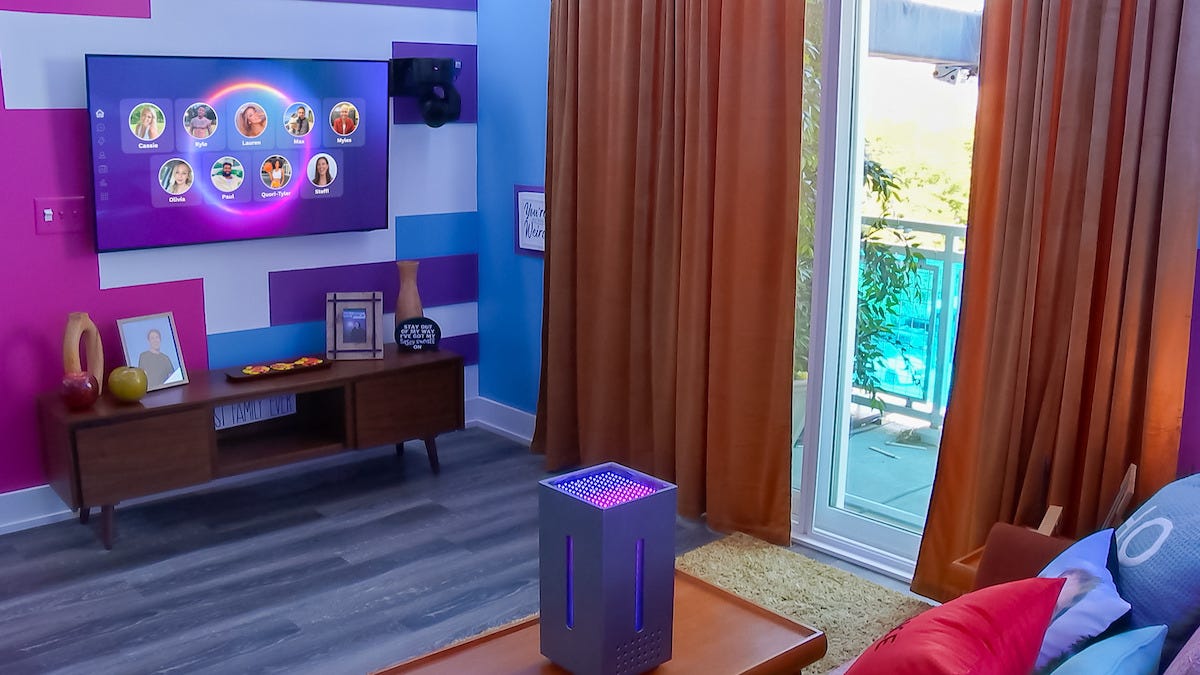 A square Alexa-looking machine on a coffee table in a living room with a TV screen featuring nine faces on it.