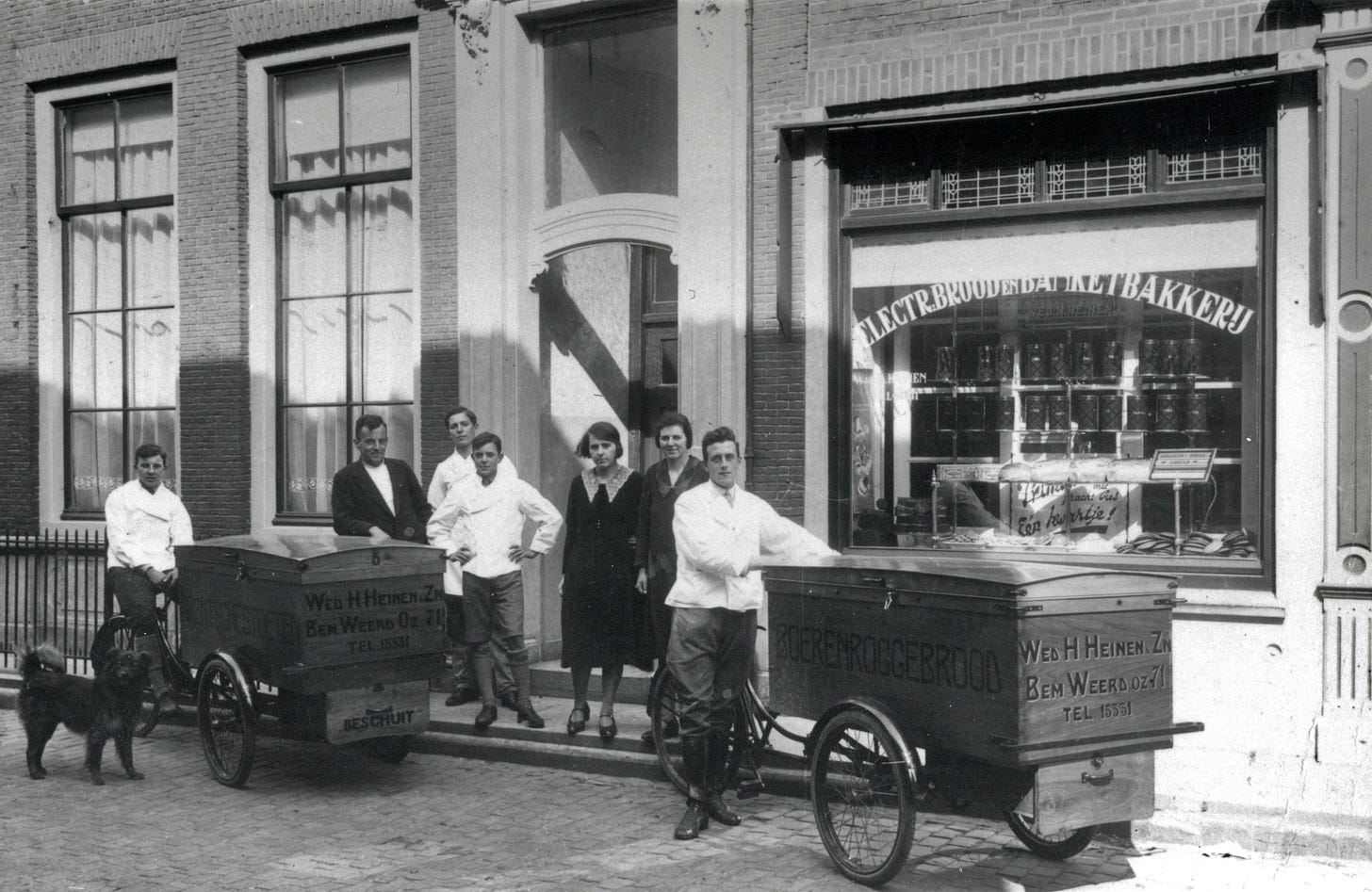 And old black and white photo showing a group of people stood infront of a bakery. There are 2 old cargobikes with them. On the cargobikes is painted text advertising the bakery