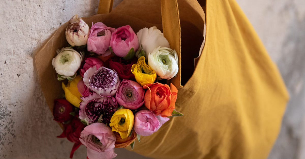 A mustard yellow tote bag filled with bright spring flowers rests against a textured white wall.