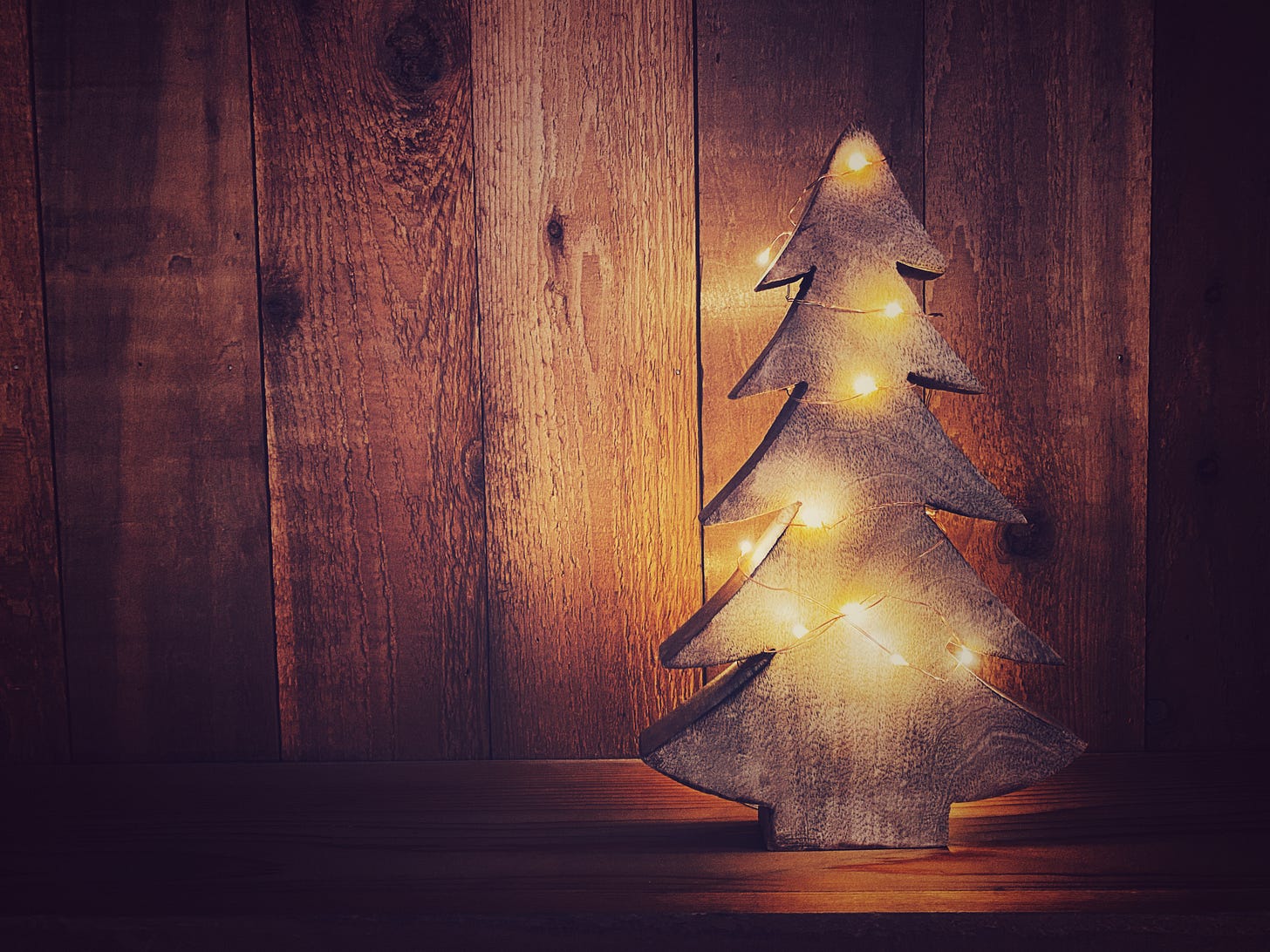 A photograph of a wooden evergreen tree strung with small, bright fairy lights standing in the dark with a rough wooden wall background.