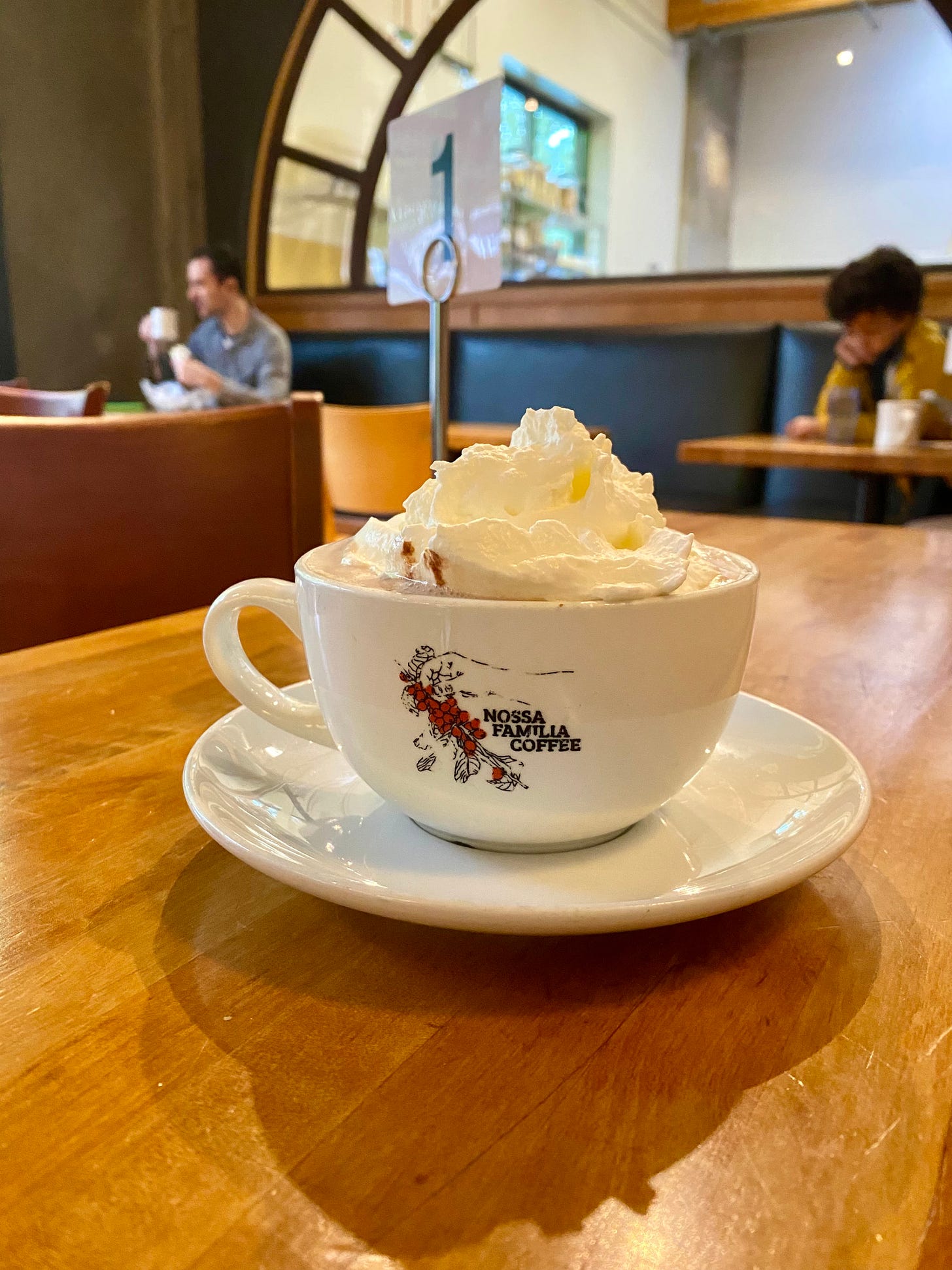 Large mug of hot chocolate with whipped cream, on a table in a coffee shop. In the background, two patrons, each sitting alone.